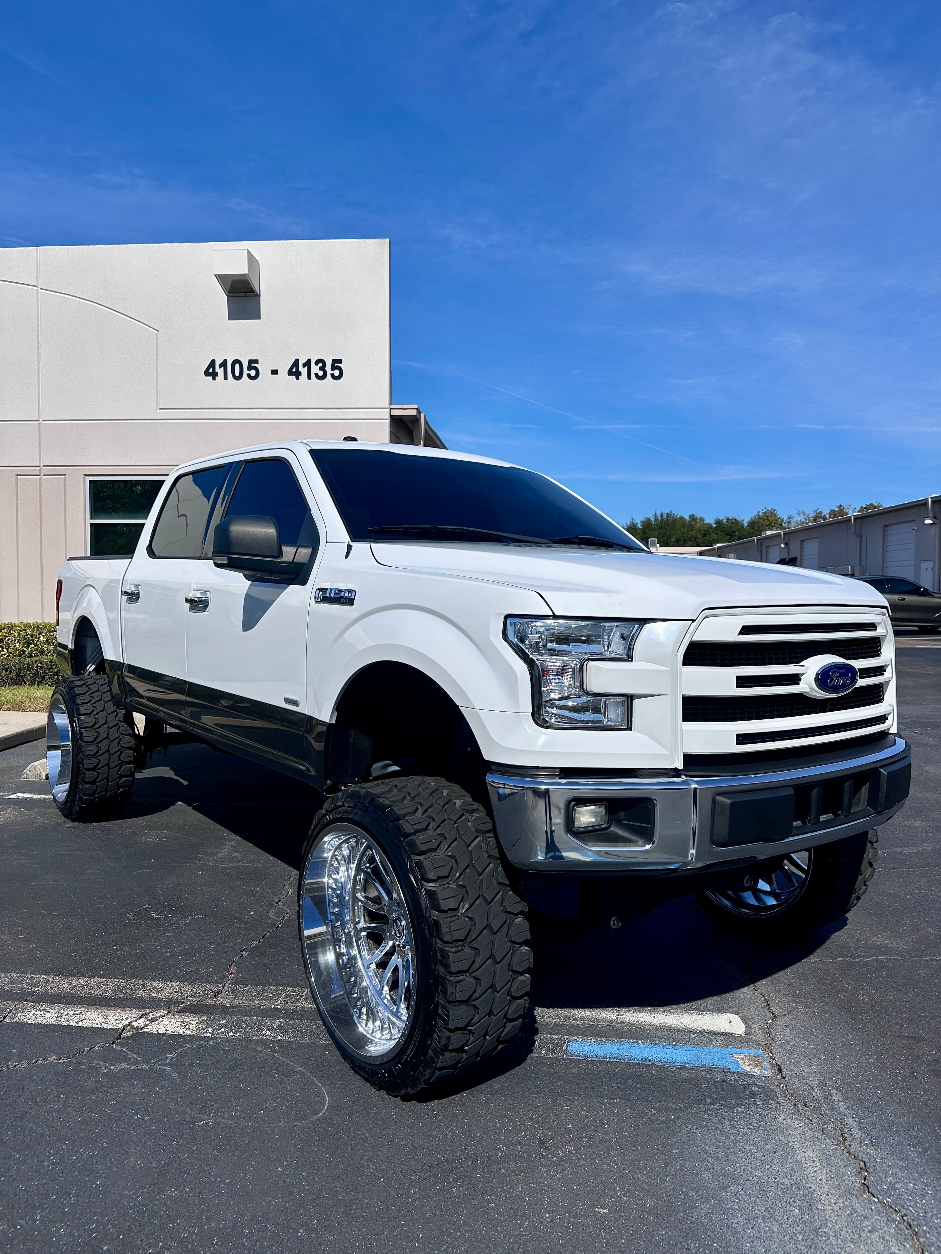 White lifted Ford F-150 pickup truck with oversized chrome wheels and off-road tires outdoors.